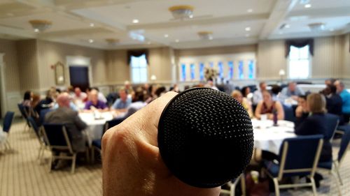 Cropped image of hand holding microphone with people sitting on chairs