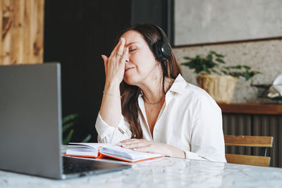 Adult tired brunette woman fifty years in headphones doing notes in daily book with laptop 