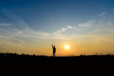 Silhouette man with hand raised standing on field against sky during sunset