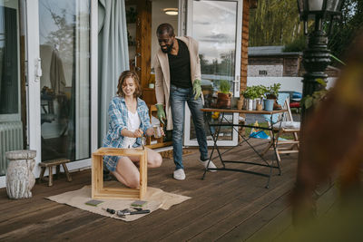 Mature man looking at woman painting furniture on porch outside house