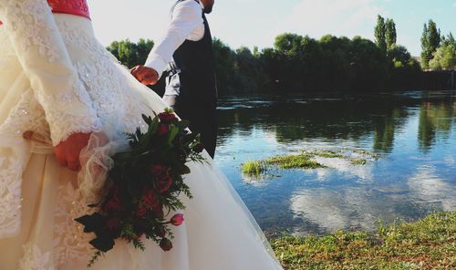 Midsection of couple holding hands while standing by lake