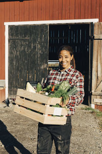 Portrait of smiling mature woman carrying crate full of vegetables with barn in background