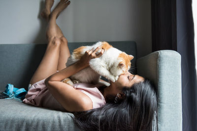 Asian young woman kiss and cuddle her yellow exotic shorthair cat on living room sofa