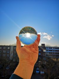 Cropped hand holding crystal ball against blue sky during sunny day