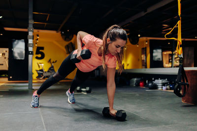 Full body focused young fit female performing push up exercise with dumbbells during intense workout in contemporary sports center with modern equipment