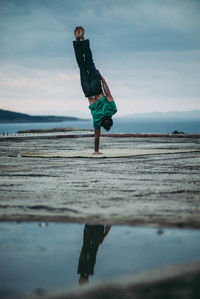 Full length of man balancing on hand against cloudy sky