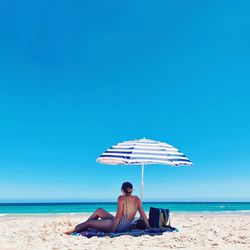 Woman sitting under parasol at beach against clear blue sky