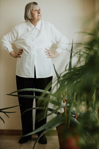 Focused female pensioner in white blouse and black trousers standing at wall looking away