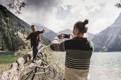 Austria, tyrol, alps, woman taking cell phone picture of man balancing on tree trunk at mountain lake