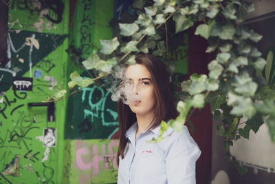 Portrait of beautiful young woman emitting smoke from mouth while standing against wall