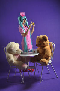 Woman wearing nurse costume holding syringe by teddy bears on chair against purple background