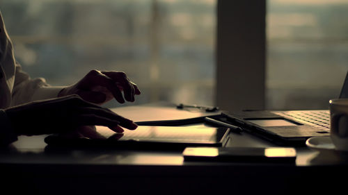 Dark silhouette of female hands. she is typing something in tablet, next is a cup, laptop on table.