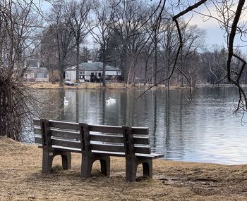 Empty bench by lake against bare trees and buildings