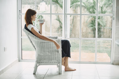 Full length of woman holding coffee cup while sitting against window