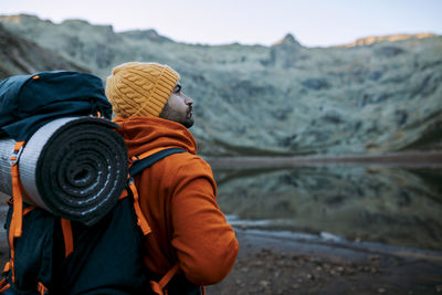 Side view of male traveler with backpack standing on shore near lake against mountainous area during trip in nature