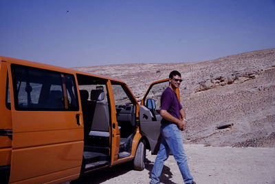 Man walking on sand by car against clear blue sky