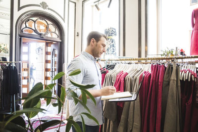 Side view of sales clerk looking at clothes rack while holding book and laptop in boutique