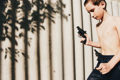 Shirtless boy holding gun by wall on sunny day