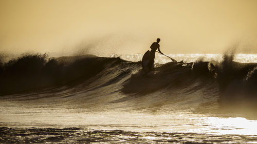 Silhouette man paddleboarding in sea against clear sky at sunset