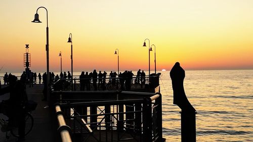 Silhouette people on street by sea against orange sky