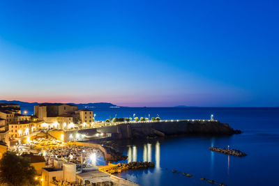 Illuminated buildings by sea against blue sky at dusk