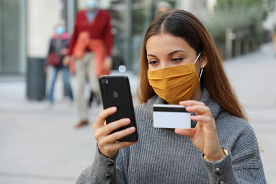 Young woman wearing mask using phone while holding credit card standing outdoors