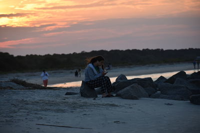 Teenage girl sitting on a rock on the beach against sky during sunset