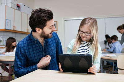 Smiling teacher looking at girl holding digital tablet at desk