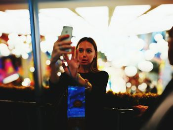 Young woman taking selfie through mobile phone while standing against illuminated lights