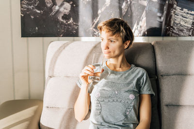 A woman sits on a couch in sunlight and holds a glass of water.
