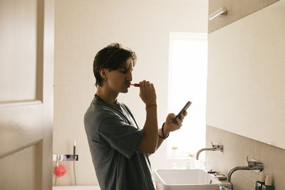 Side view of young man using smart phone while brushing teeth in bathroom at home