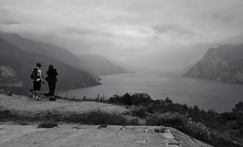 Rear view of couple standing on mountain while looking at lake amidst sky