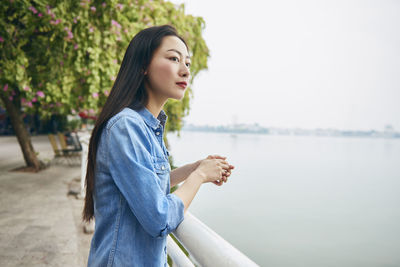 Side view of young woman looking away standing by railing against river