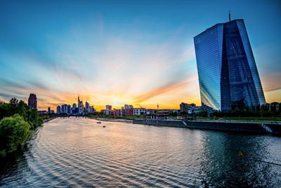 View of modern buildings by river against sky during sunset