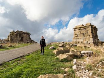 Full length of woman standing on grassy field at old ruins against cloudy sky