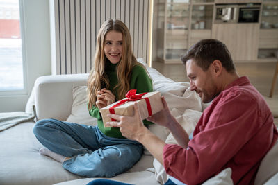 Intrigued man sitting on sofa, trying to unwrap giftbox, and glad woman looking at him with love