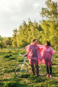 Young couple in pink raincoat standing with bicycle on field against sky