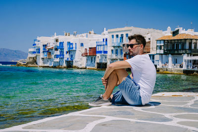 Man sitting on building by sea against sky