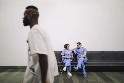 Happy male and female medical experts talking while sitting on chair near wall in hospital