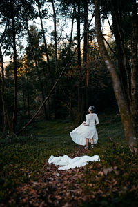 Full body of anonymous female wearing white dress standing near green trees in nature on evening time