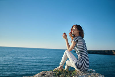 Woman sitting by sea against clear blue sky