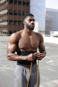 Portrait of shirtless man holding camera while standing against built structure