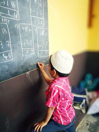 Side view of boy writing on blackboard while standing in classroom