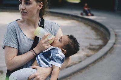 Mother feeding baby boy from bottle while standing at park