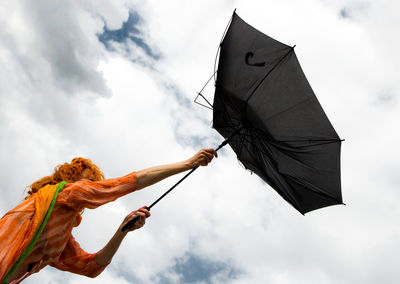 Low angle view of mature woman holding damaged umbrella against cloudy sky