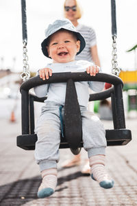 Mother pushing her infant baby boy child on a swing on playground outdoors.