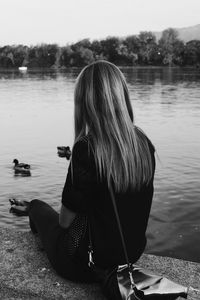 Rear view of woman sitting on retaining wall while looking at lake