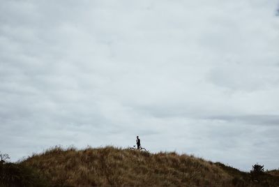 Man with bicycle standing on grassy hill against cloudy sky