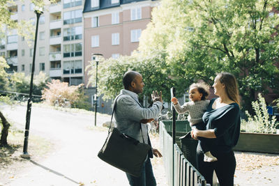 Mother carrying daughter waving father at park in city