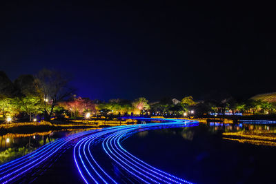 Illuminated light trails on river by city against sky at night